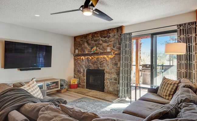 Living room with stone fireplace, wall-mounted TV, sectional sofa, and sliding glass doors opening to a deck with mountain views