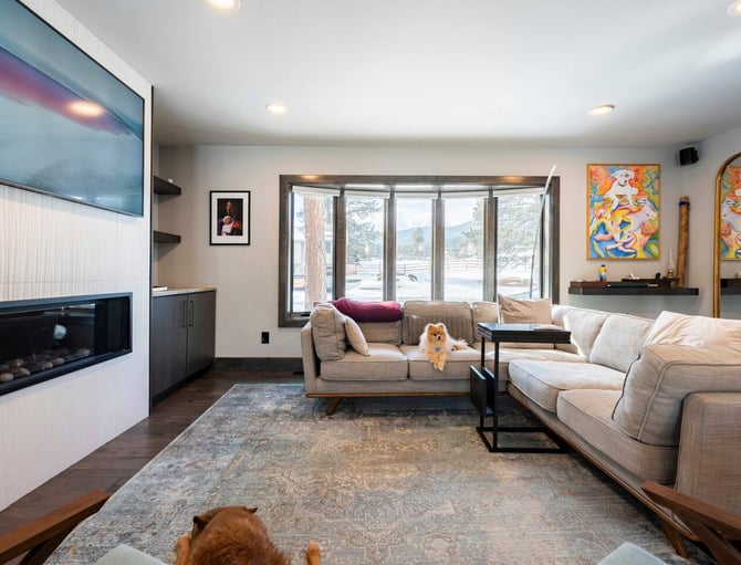 Modern living room with beige sectional sofa, fireplace, wall-mounted TV, large windows overlooking mountains, and two small dogs on furniture